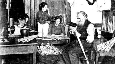Eine fünfköpfige Familie sitzt am Tisch ihrer Einzimmer-Wohnung und fertigt Papptröten für Jahrmärkte. Auch ein Kind hilft bei der Heimarbeit mit.