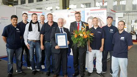 Dieter Mießen hält die Urkunde, Thomas Frisch den Blumenstrauß, und alle Mitarbeiter freuen sich mit ihnen.