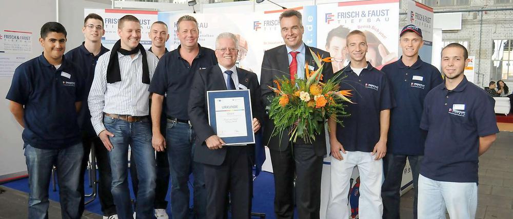 Dieter Mießen hält die Urkunde, Thomas Frisch den Blumenstrauß, und alle Mitarbeiter freuen sich mit ihnen.