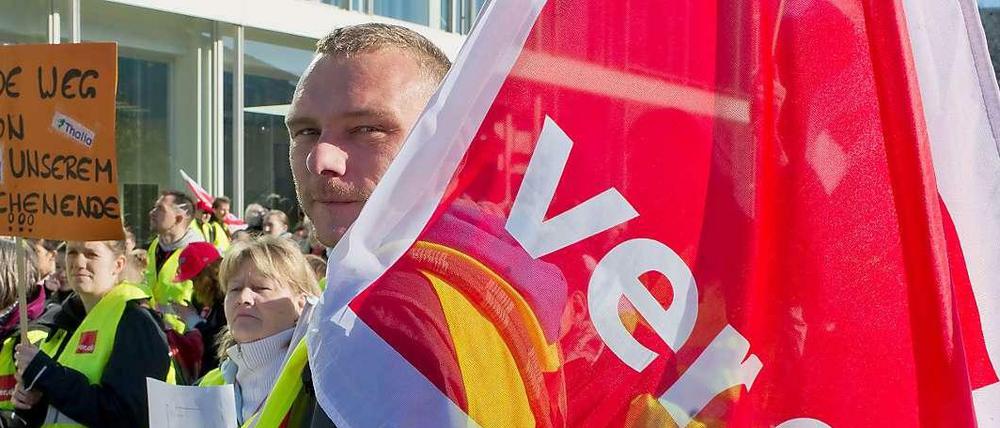 Ein Verdi-Mitglied trägt eine Fahne seiner Gewerkschaft auf der Schulter und steht zusammen mit anderen Beschäftigten vor einem Gebäude.