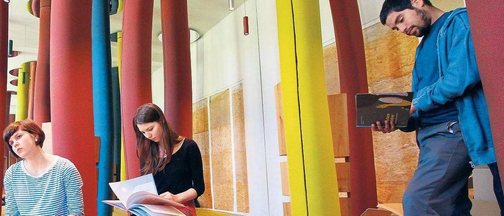 In einem künstlerisch gestalteten Raum sitzen oder stehen drei junge Menschen und lesen in Bilderbüchern.