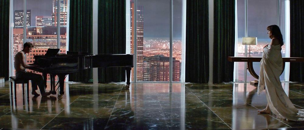 Ein Mann am Klavier und eine Frau nur bedeckt von einem Lacken vor einer Skyline