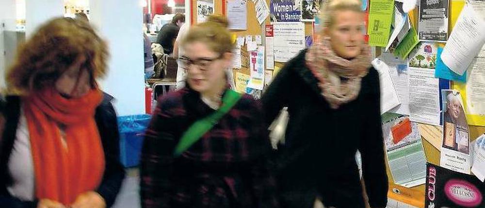 Drei Studierende laufen an einem Schwarzen Brett in der Uni vorbei.