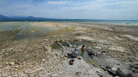 Ein Mann sitzt neben einem Kind am Gardasee, wo Steine durch den niedrigen Wasserstand freigelegt sind. 