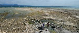 Ein Mann sitzt neben einem Kind am Gardasee, wo Steine durch den niedrigen Wasserstand freigelegt sind. 