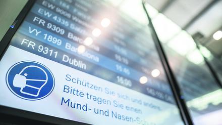 Auf einer Anzeigetafel im Flughafen steht der Hinweis „Schützen Sie sich und andere. Bitte tragen Sie einen Mund- und Nasen-Schutz“.