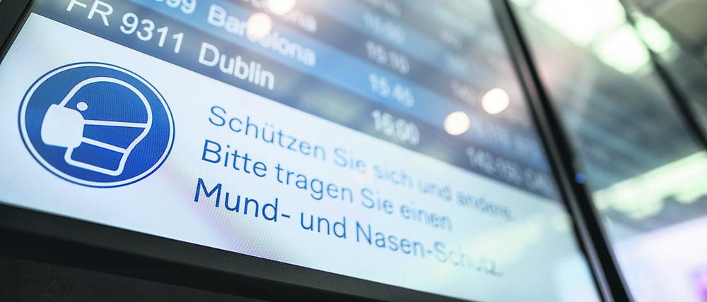 Auf einer Anzeigetafel im Flughafen steht der Hinweis „Schützen Sie sich und andere. Bitte tragen Sie einen Mund- und Nasen-Schutz“.