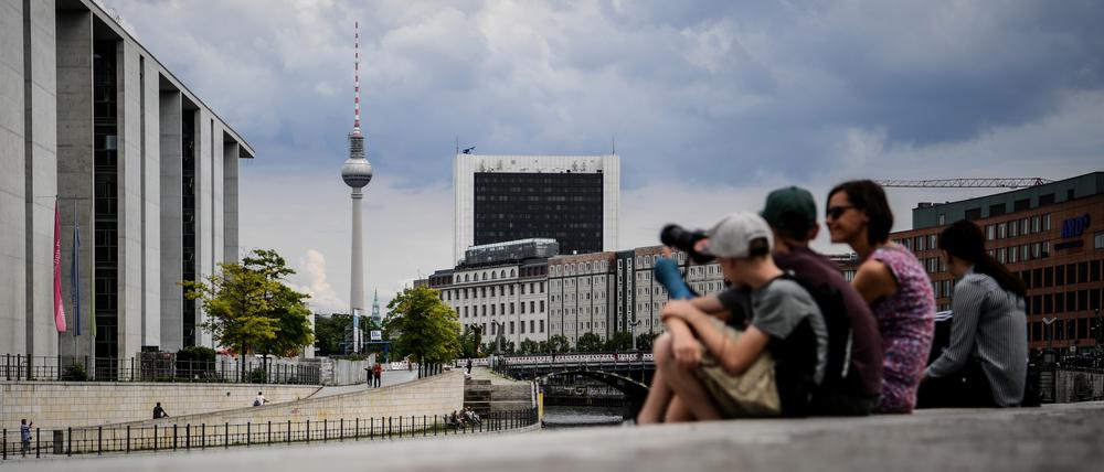 Touristen sitzen am Spreeufer in der Nähe des Bundestags. Im Hintergrund ragt der Fernsehturm am Alexanderplatz in die Höhe.