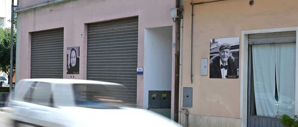 An der Fassade der Häuser hängen Schwarz-Weiß-Fotos der Hundertjährigen aus dem Dorf mit deren Namen und Geburtsjahr. Rechts auf dem Bild ist Vittorio Lai zu sehen.  Foto: dpa/Johannes Neudecker