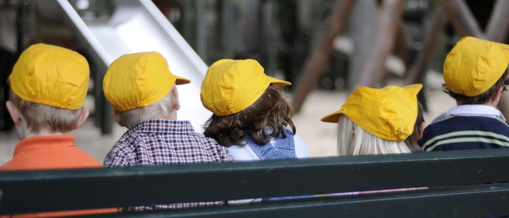 Kleine Schulkinder mit gelben Mützen sitzen in Berlin vor einem Spielplatz auf einer Bank.