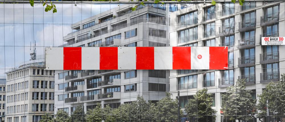Ein rot-weiße Absperrung ist an einem Foto mit Wohn- und Geschäftshäusern in Berlin angebracht über dem die Zweige von Bäumen zu sehen sind. Das Plakat mit dem Foto dient an einem Bauzaun als Sichtschutz.