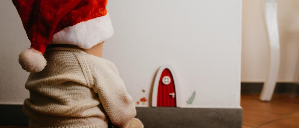Ein Kind mit Weihnachtsmannmütze sitzt vor einer kleinen Wichteltür in der Wand.
