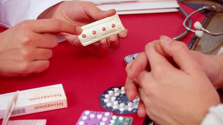 Frauenhand hält Pille für danach in Hand am Schreibtisch, Männerhände gegenüber, andere Antibabypillen auf Tisch, Gespräch, Beratung, Arztpraxis.