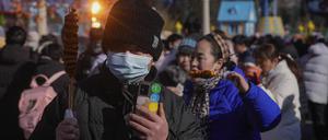 24.01.2023, China, Peking: Ein Mann mit einer Gesichtsmaske hält gegrillten Tintenfisch, während die Besucher eines Tempelmarktes während des einwöchigen Neujahrsfestes vorbeischauen. Foto: Andy Wong/AP/dpa +++ dpa-Bildfunk +++