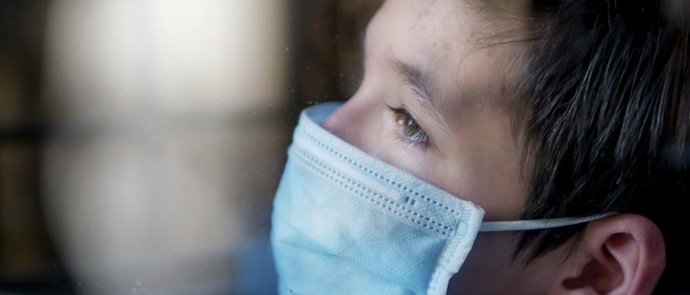 Trauriger Junge mit medizinischer Gesichtsmaske hinter einem Fenster während der Ausganssperre