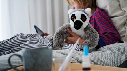 Fieberthermometer, Nasenspray und eine Tasse Tee stehen vor einem Bett, in dem ein Kind mit Kuscheltier auf ein Tablet guckt.