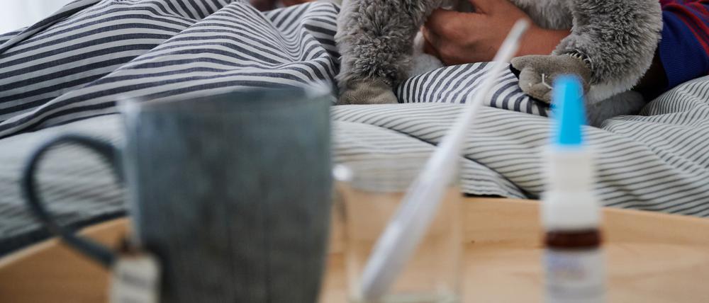 Fieberthermometer, Nasenspray und eine Tasse Tee stehen vor einem Bett, in dem ein Kind mit Kuscheltier auf ein Tablet guckt.