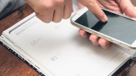 Eine Person schaut etwas in ihrem Smartphone nach und hat ihren Kalender offen vor sich liegen.