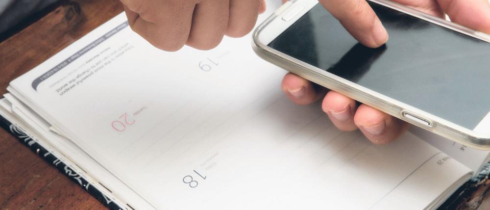 Eine Person schaut etwas in ihrem Smartphone nach und hat ihren Kalender offen vor sich liegen.
