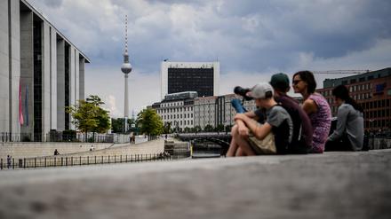 Touristen sitzen am Spreeufer in der Nähe des Bundestags. Im Hintergrund ragt der Fernsehturm am Alexanderplatz in die Höhe. 