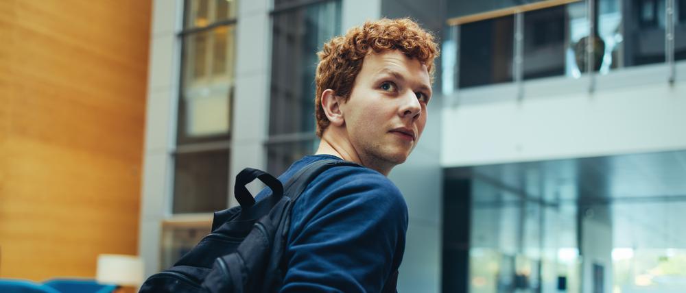 College student carrying his bag and laptop in campus. Young man turning back over his shoulder and walking in college campus.