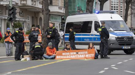 Aktivisten der Gruppe „Letzte Generation“ haben sich auf der Neuen Kantstraße auf einer Kreuzung festgeklebt. Die Polizei sichert den Bereich, während eine Polizistin begonnen hat, die Aktivisten von der Straße zu lösen.