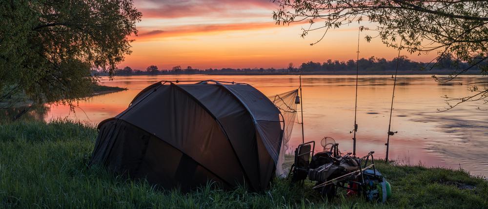 Farbenprächtig leuchtet der Morgenhimmel kurz vor Sonnenaufgang über dem deutsch-polnischen Grenzfluss Oder am Rande des Oderbruchs in Ostbrandenburg. Am Ufer stehen ein Zelt und Angeln. Auch Kinder mögen dieses Hobby – für sie gibt es in Berlin und Brandenburg unterschiedliche Regelungen.