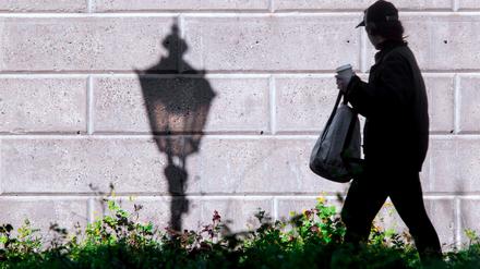 Eine Straßenlaterne wirft im Licht der aufgehenden Sonne einen Schatten an eine Wand, während eine Frau vorbeigeht.