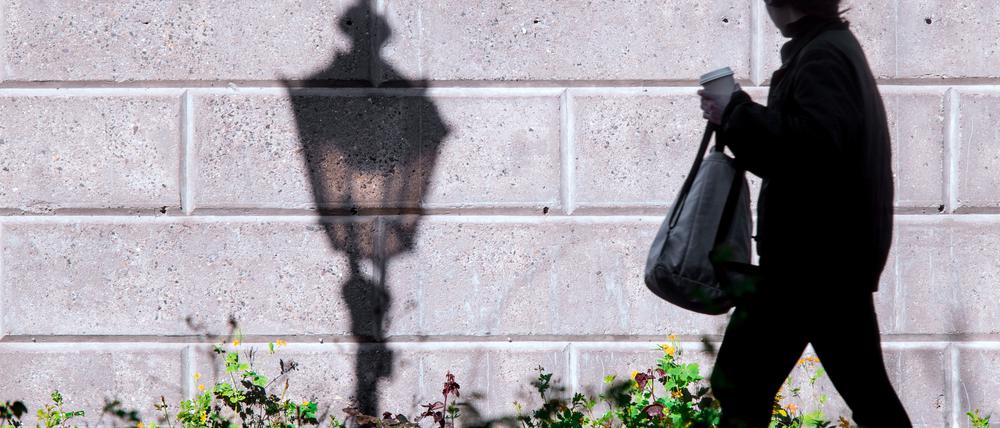 Eine Straßenlaterne wirft im Licht der aufgehenden Sonne einen Schatten an eine Wand, während eine Frau vorbeigeht.