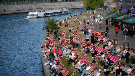 Ein Ausflugsschiff fährt bei sommerlichen Temperaturen auf der Spree, während am Ufer Touristen auf Liegen die Sonne genießen.