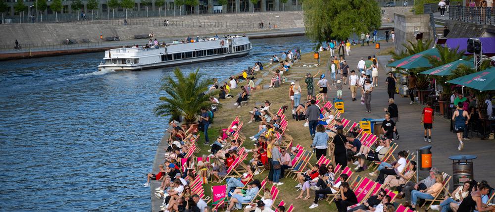 Ein Ausflugsschiff fährt bei sommerlichen Temperaturen auf der Spree, während am Ufer Touristen auf Liegen die Sonne genießen.