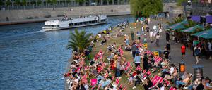 Ein Ausflugsschiff fährt bei sommerlichen Temperaturen auf der Spree, während am Ufer Touristen auf Liegen die Sonne genießen.