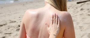 Eine Person cremt einer blonden Frau am Strand den Rücken mit Sonnencreme ein.