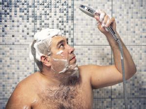 Ein Mann steht mit Shampoo im Haar unter der Dusche und blickt unsicher auf den Duschkopf in seiner Hand.