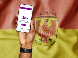 Montage einer Hand die ein Mobiltelefon hält auf dessen Bildschirm die Internetseite des Votomats abgebildet ist – vor einer Potsdam Flagge.