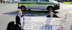 Klimaaktivisten der Gruppe Letzte Generation sitzen mit Masken von Bundeskanzler Scholz (l) und Wirtschaftsminister Habeck bei einer Sitzblockade auf einer Kreuzung vor einem Polizeiauto. Die Aktivisten halten ein Banner mit der Aufschrift „Wir brechen das Gesetz!“