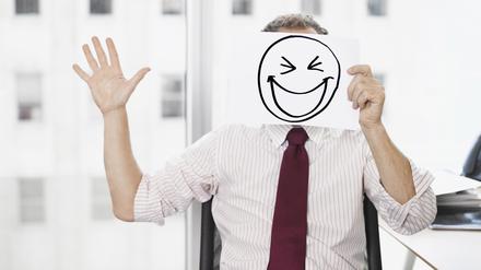 Ein Geschäftsmann hält ein Papier mit einem gemalten, breit grinsenden Smiley vor seinem Gesicht.