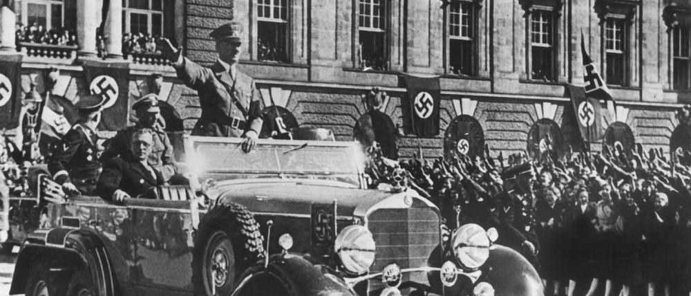 In einem Auto stehend grüßt Adolf Hitler die ihm zujubelnde Menschenmenge in Wien.