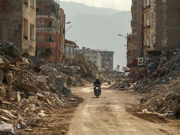 A man rides a motorcycle along the rubble of destroyed buildings. 