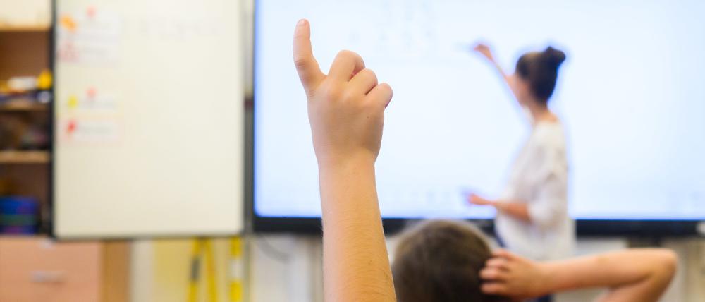 Ein Schüler meldet sich per Handzeichen während eine Lehrerin auf eine digitale Schultafel im Klassenraum einer 4. Klasse einer Grundschule in der Region Hannover schreibt. 