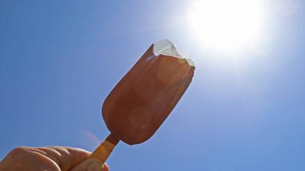 Die Sonne strahlt am Mittag vom blauen wolkenlosen Himmel. Davor ein Eis am Stiel mit Schokolade (Archivbild).