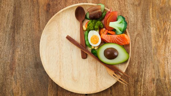Gemüse und ein Ei liegen auf einem Holzteller, auf dem Löffel und Gabel wie die zwei Zeiger einer Uhr angeordnet sind. (Symbolbild fürs Intervallfasten)