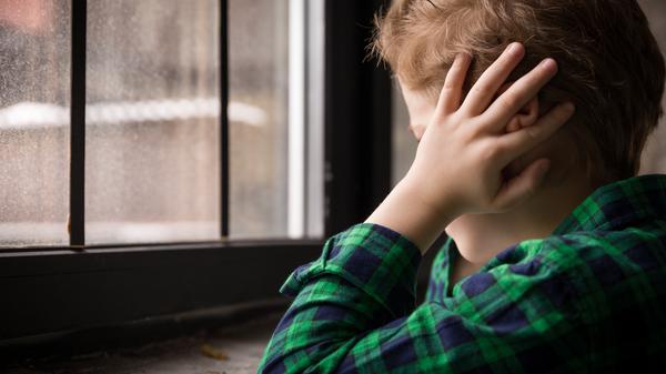 Ein kleiner Junge sitzt traurig vor dem Fenster und hält sich die Ohren zu.
