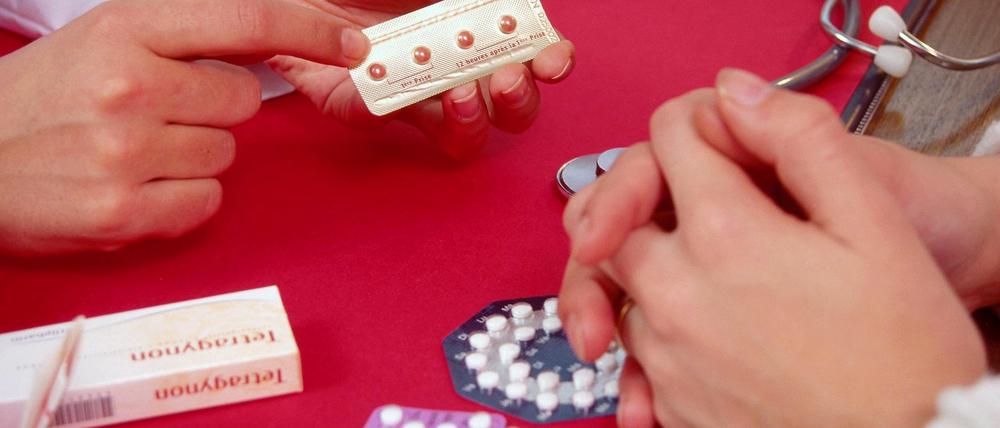 Frauenhand hält Pille für danach in Hand am Schreibtisch, Männerhände gegenüber, andere Antibabypillen auf Tisch, Gespräch, Beratung, Arztpraxis.