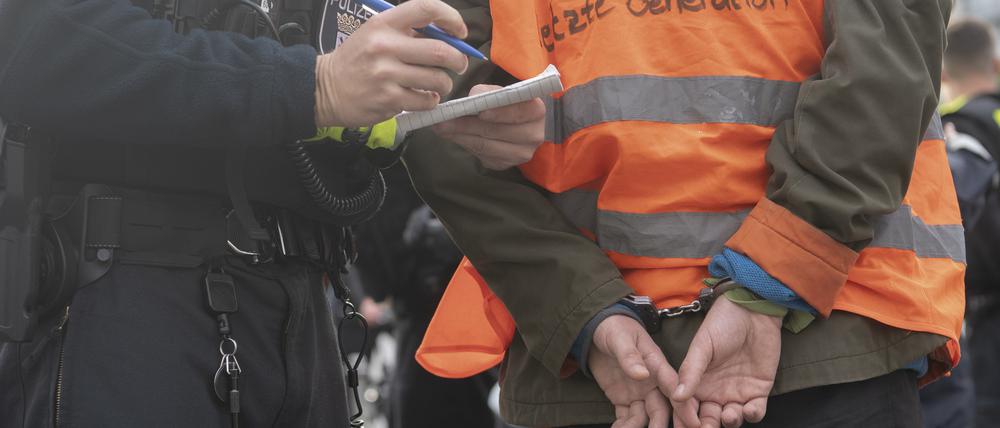 Ein Aktivist der Klimaschutz-Gruppe Letzte Generation steht am Potsdamer Platz in Handschellen neben einem Polizeibeamten.