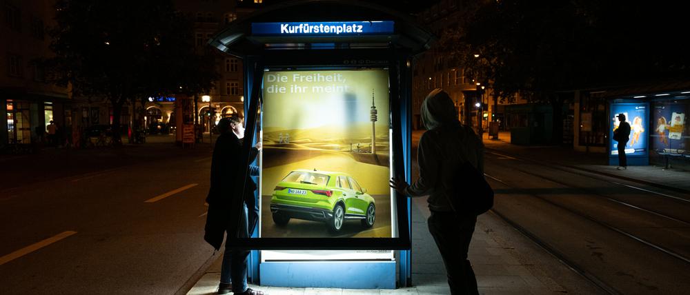 07.09.2023, Bayern, München: Aktivisten der Nichtregierungsorganisation Attac bringen als Protest gegen die aktuell in München laufende Messe IAA Mobility kritische Persiflagen von Autowerbung an einem Trambahnhäuschen an.