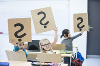Schüler und Schülerinnen sitzen in einer Klasse und halten Pappschilder mit Fragezeichen hoch.