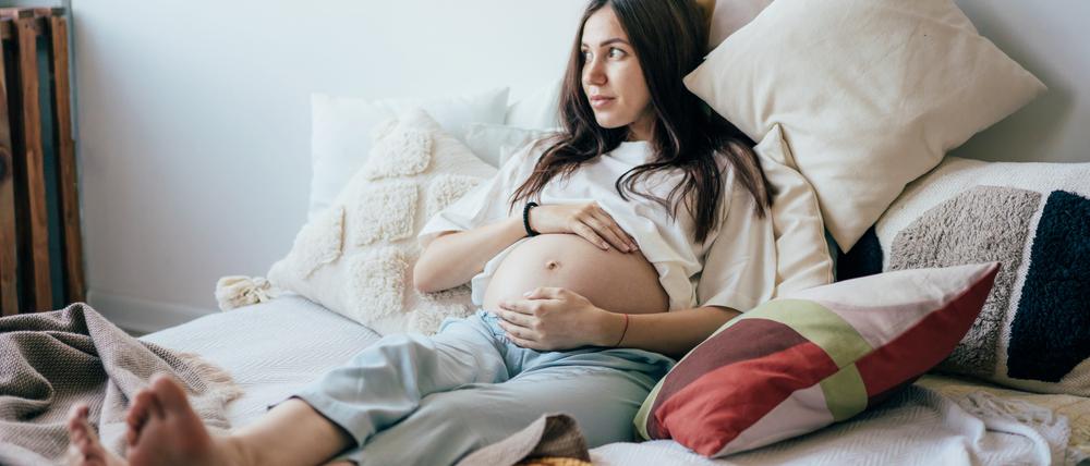 Eine junge schwangere Frau liegt in ihrem Bett und hält ihren Bauch.