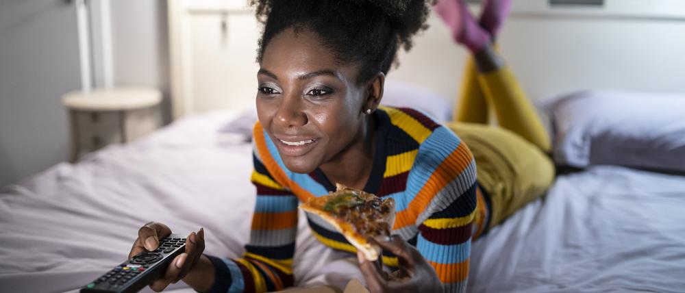 Eine Frau isst Pizza im Bett und hat eine Fernbedienung in der Hand