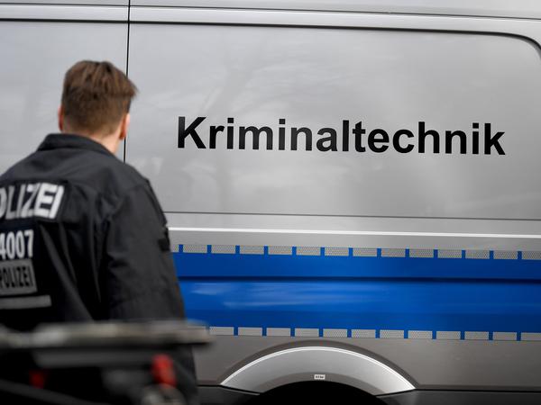 Ein Berliner Polizist steht neben einem Fahrzeug der Kriminaltechnik mit Berliner Kennzeichnen, das an einem Waldweg parkt.